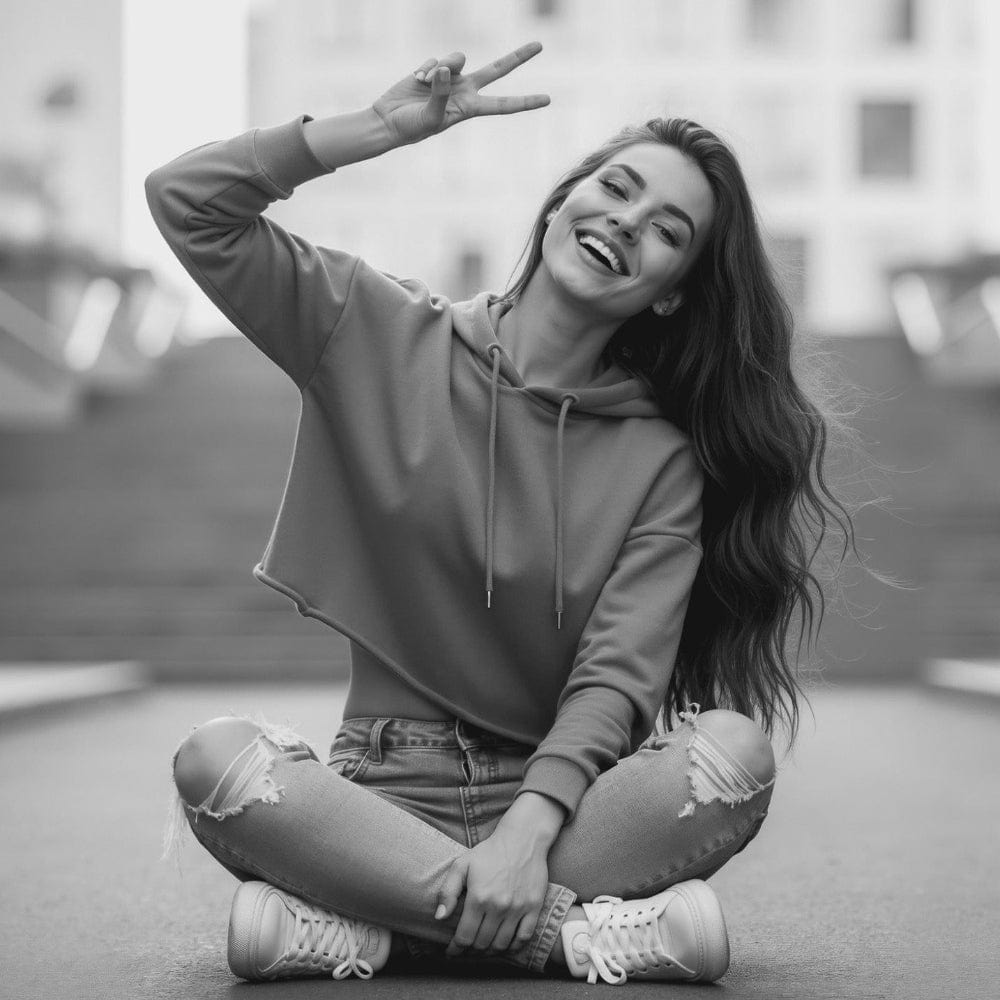 Black and white photo of a woman sitting on the ground with a peace sign, wearing a hoodie and ripped jeans.